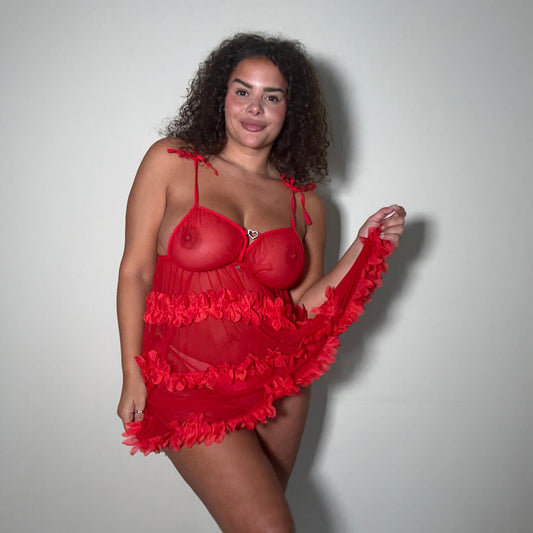 A woman modeling a red sheer lingerie set  from Freak Geek Lingerie. with ruffled trim, standing against a plain light gray wall.