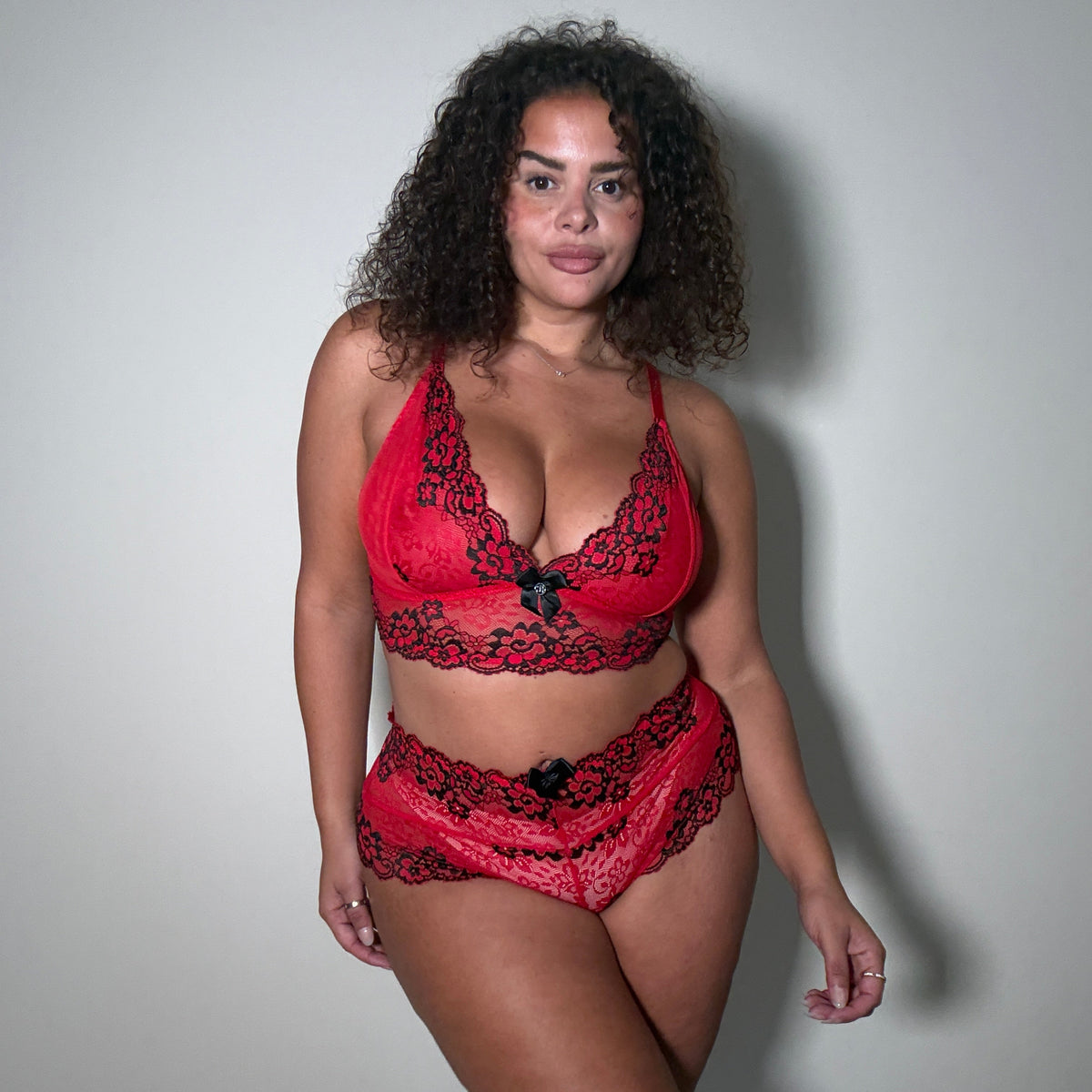 A woman modeling a red and black lace lingerie set consisting of a bralette and matching panties, standing against a plain light gray wall.
